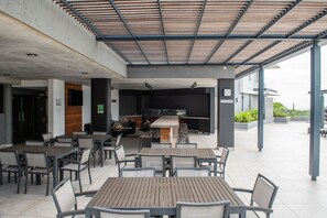 An outdoor patio area with multiple tables and chairs, a wooden pergola overhead, and a bar area in the background.