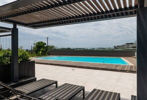 A rooftop pool area with a wooden pergola, lounge chairs, and a view of the city skyline.