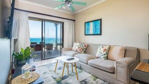 A living room with a sofa, coffee table, and a ceiling fan. There is a large window with a view of the ocean.