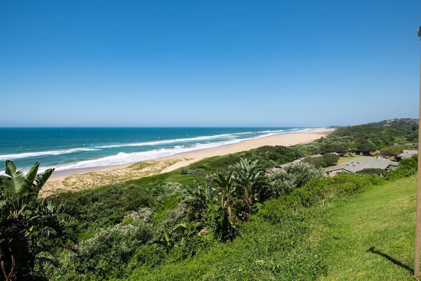 Paisaje costero con playa, vegetación y edificios a lo lejos.