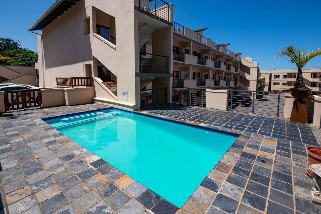 A swimming pool with a tiled deck, a multi-story building with balconies, and a clear blue sky.