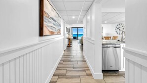 A hallway with a view to a dining area, a kitchen with a dishwasher, and a clock on the wall. - Edgewater Tower 3-104 (Panama City Beach)