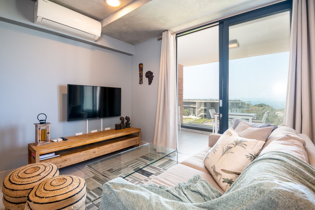 A modern living room with a flat-screen TV, wooden entertainment unit, and a large sliding glass door leading to a balcony with a view.