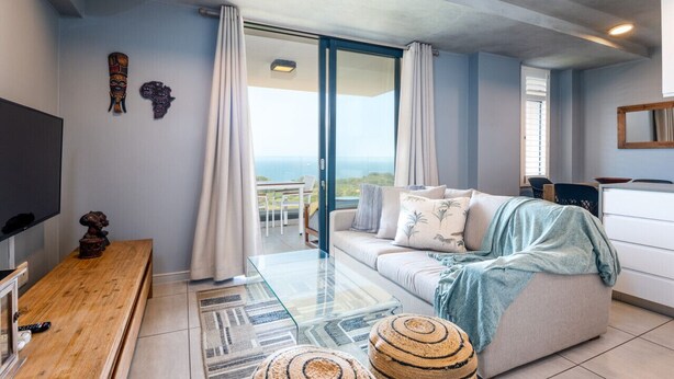 Ein modernes Wohnzimmer mit einer Couch, einem Couchtisch und einem Fernseher. Es gibt einen Balkon mit Vorhängen und einer Aussicht.