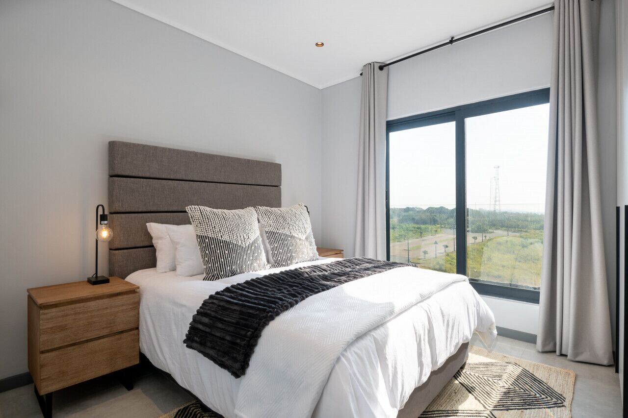 A modern bedroom with a large bed, a nightstand, a bedside lamp, and a view of a landscape through a large window.