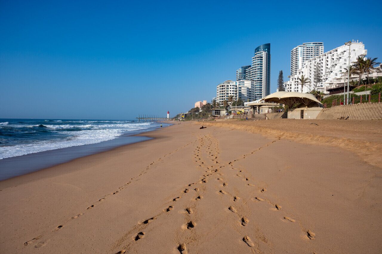 Una playa con huellas que conducen al agua, un faro y edificios modernos al fondo.