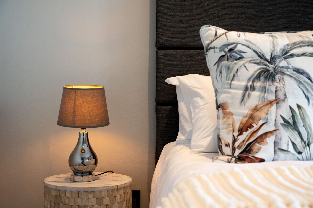A bed with a white comforter and a pillow with a tropical leaf pattern, a round wooden nightstand with a lamp, and a dark upholstered headboard.