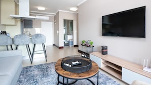 Ein modernes Wohnzimmer mit rundem Couchtisch, grauen Barhocker, einem Flachbildfernseher und einer Küchenzeile im Hintergrund.