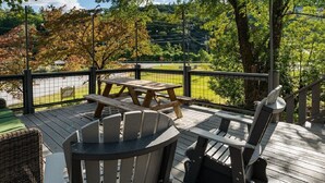 A wooden deck with a picnic table and Adirondack chairs, surrounded by a metal fence and trees.