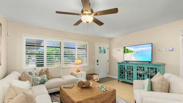 A living room with a ceiling fan, a television, and a view of the beach through the windows. - SwimSpa Oasis Backyard Beach 1.5 mi Monthly Stays (Naples)