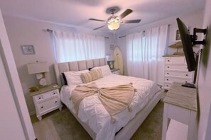 Et soveværelse med seng, natbord, kommode, loftventilator og et vindue med gardiner.