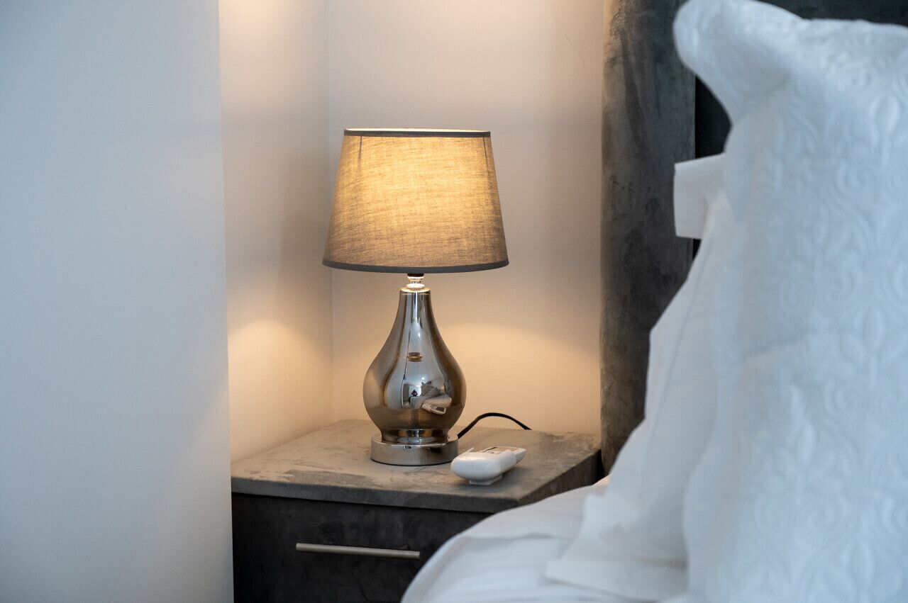 A bedside table with a lamp, a remote control, and a bed with white bedding.