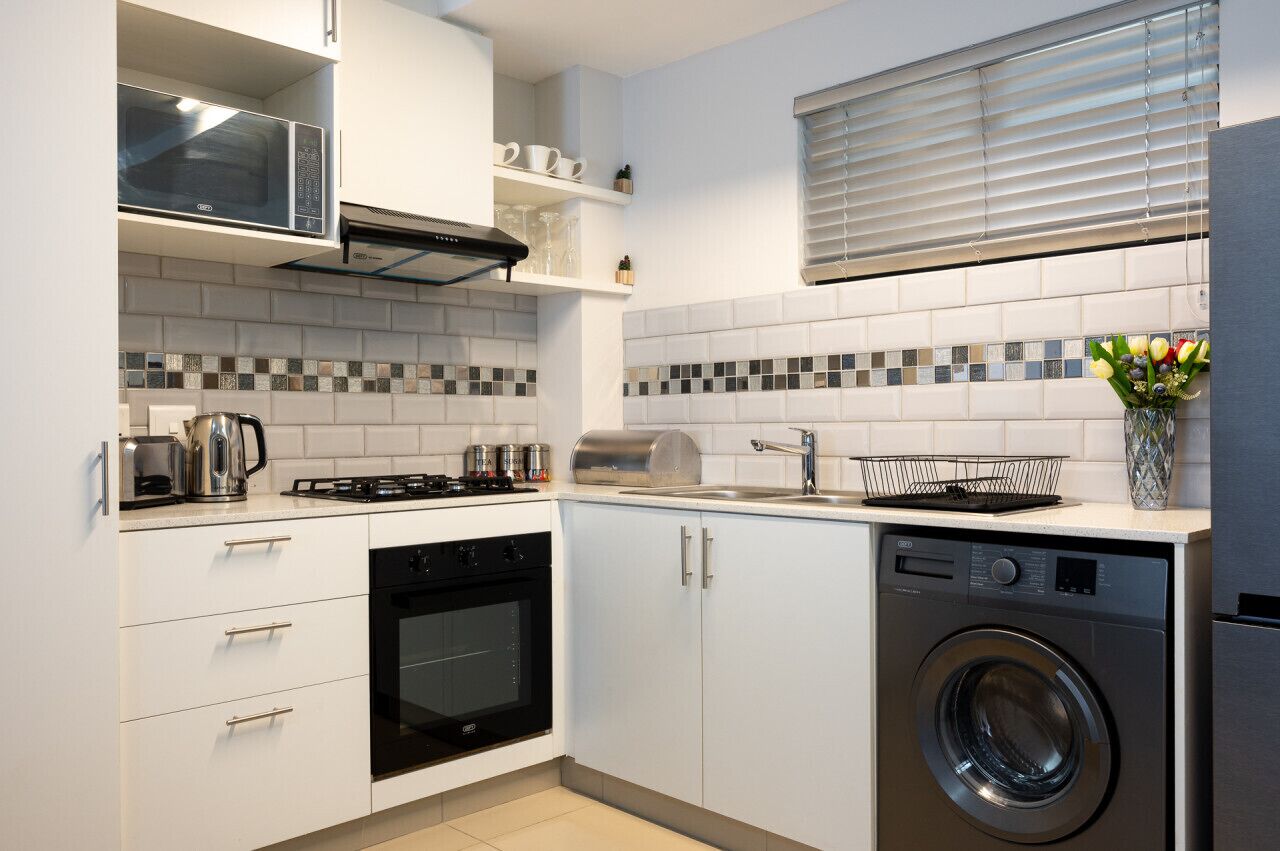 A modern kitchen with white cabinets, a built-in oven, a microwave, a dishwasher, and a window with blinds.