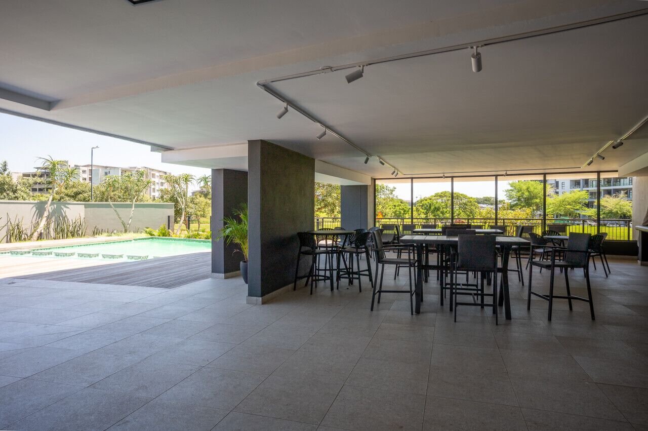 Un vaste espace extérieur aménagé pour les repas, doté de mobilier moderne et de grandes fenêtres offrant une vue sur la verdure et les bâtiments.