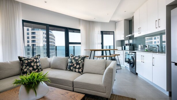 Un appartement moderne doté d’un espace de vie comprenant un canapé, une table basse agrémentée d’une plante et une table à manger. La cuisine est équipée d’un lave-linge et de placards.