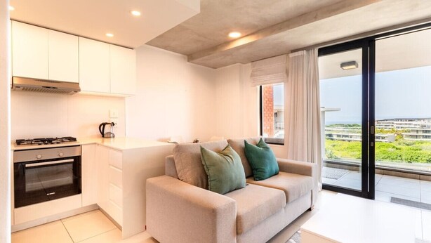 Una cucina moderna con mobili bianchi e forno integrato, un divano beige con cuscini e un'ampia porta scorrevole in vetro che dà su un balcone con vista sulla città.