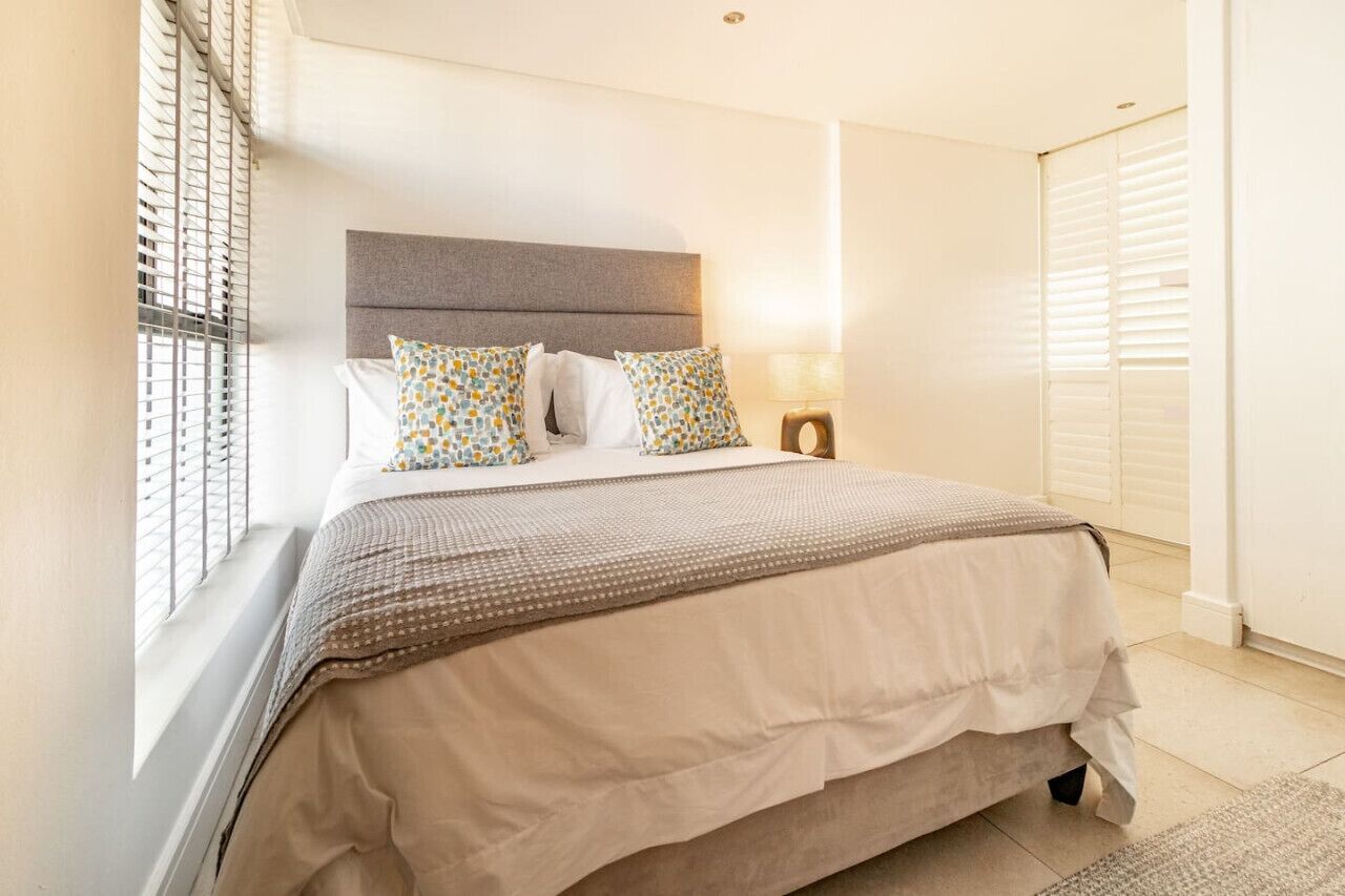 Una cama bien hecha con un cabecero gris, almohadas blancas y estampadas, y una colcha color beige.