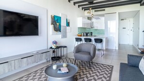 Una sala de estar moderna con un televisor grande, un sillón gris, una mesa de centro y una cocina al fondo.
