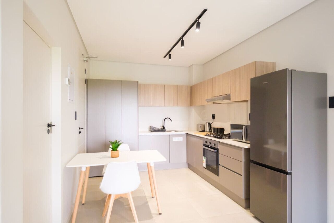 A modern kitchen with light wood cabinets, a stainless steel refrigerator, a white countertop, and a small dining table with a plant.