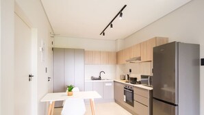 A modern kitchen with light wood cabinets, a stainless steel refrigerator, a white countertop, and a small dining table with a plant.