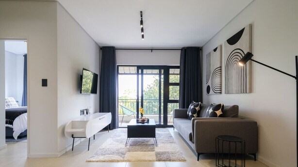A modern living room with a sofa, a coffee table, and a TV mounted on the wall. There is a large window with curtains and a balcony visible through the window.