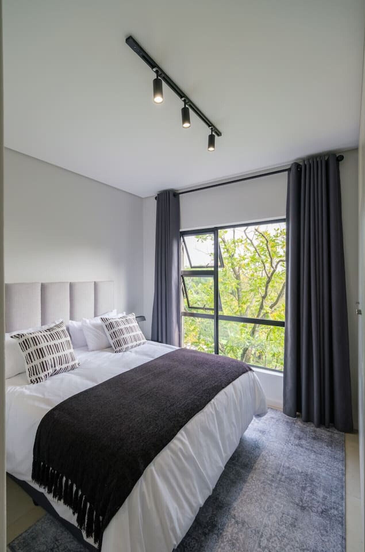 A modern bedroom with a large bed, grey curtains, and a view of trees through the window.