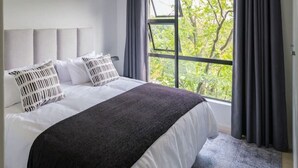 A modern bedroom with a large bed, grey curtains, and a view of trees through the window.