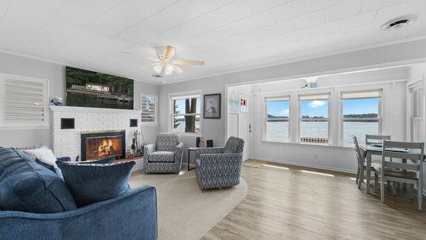 A spacious living room with a fireplace, comfortable seating, large windows with a view of the water, and a ceiling fan.