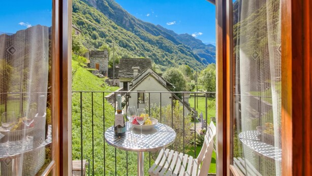 A balcony with a table set for two, a bottle, and a bowl of fruit. - Ca Contina in Val Verzasca (Sonogno)