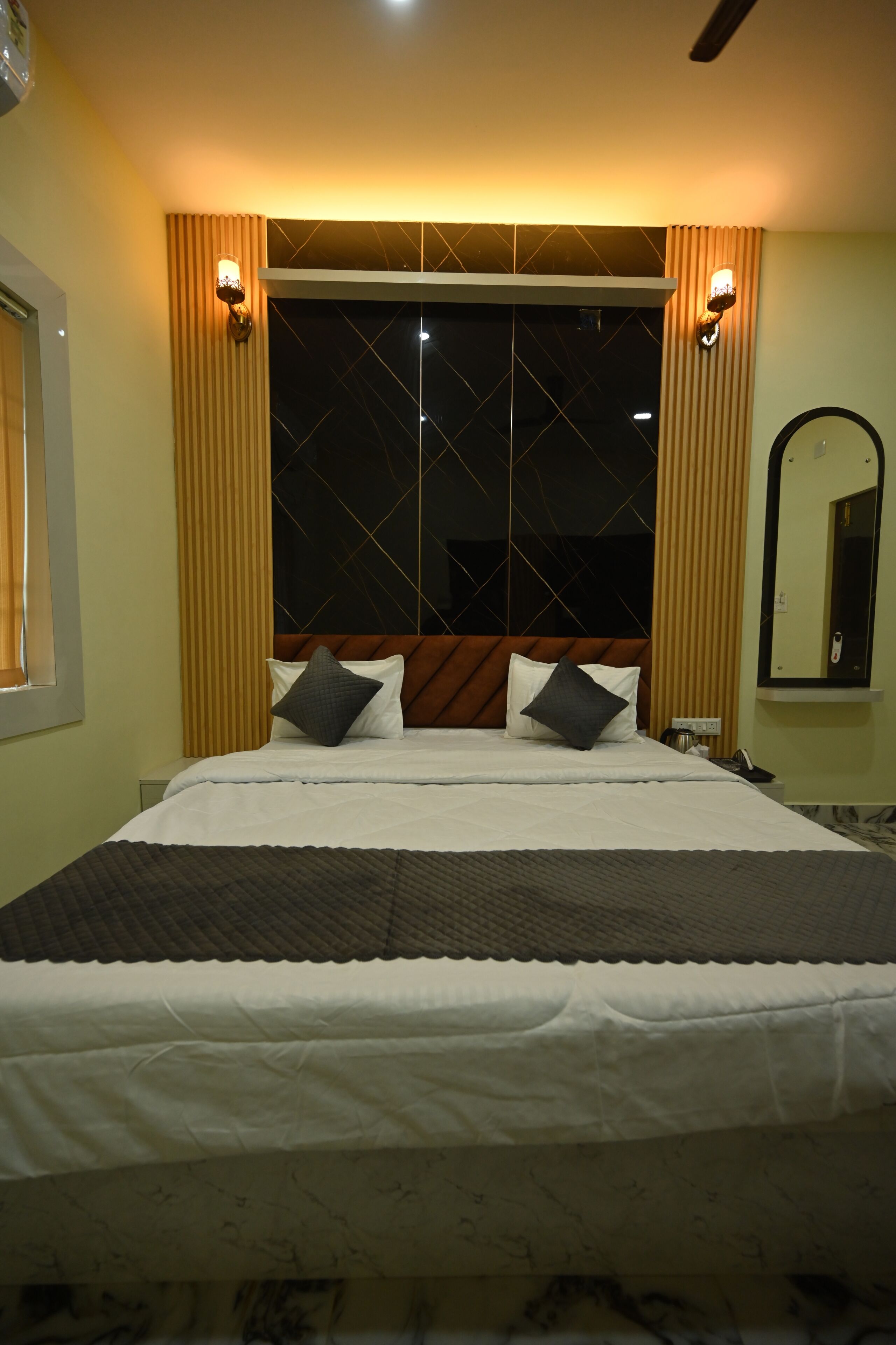A hotel room with a large bed, a headboard with a geometric pattern, two wall-mounted light fixtures, a mirror, and a window with curtains.