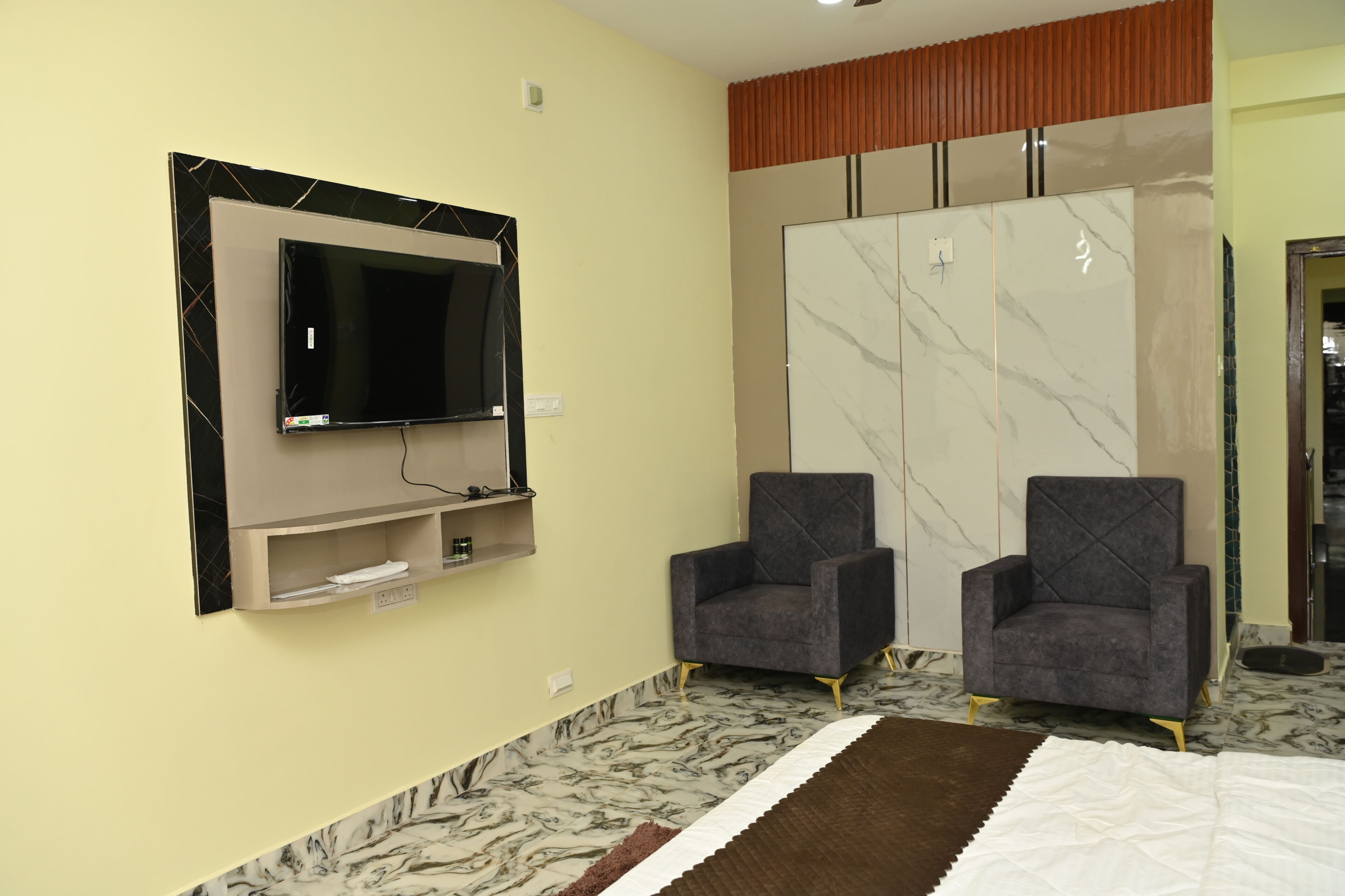 Una habitación de hotel con una televisión de pantalla plana montada en la pared, dos sillones, un sofá, un suelo con diseño de mármol y un armario corredero.