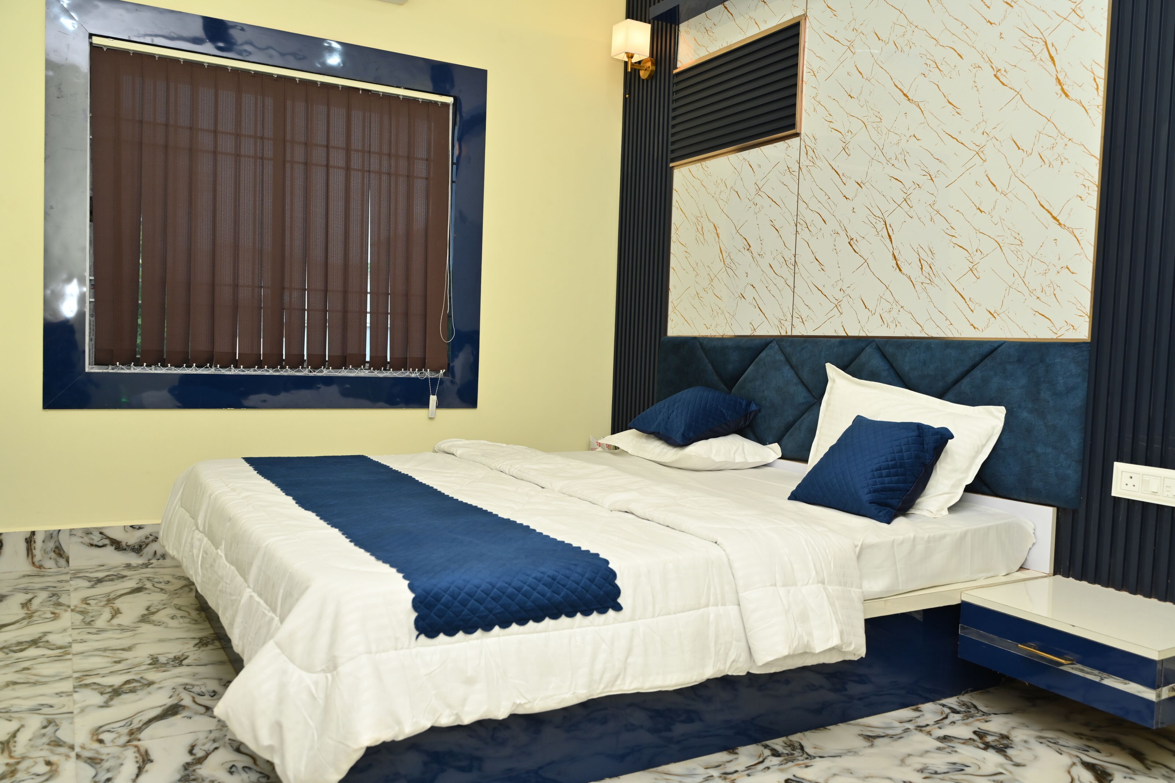 A bedroom with a bed, a nightstand, a wall-mounted lamp, a window with blinds, and a marble-patterned floor.