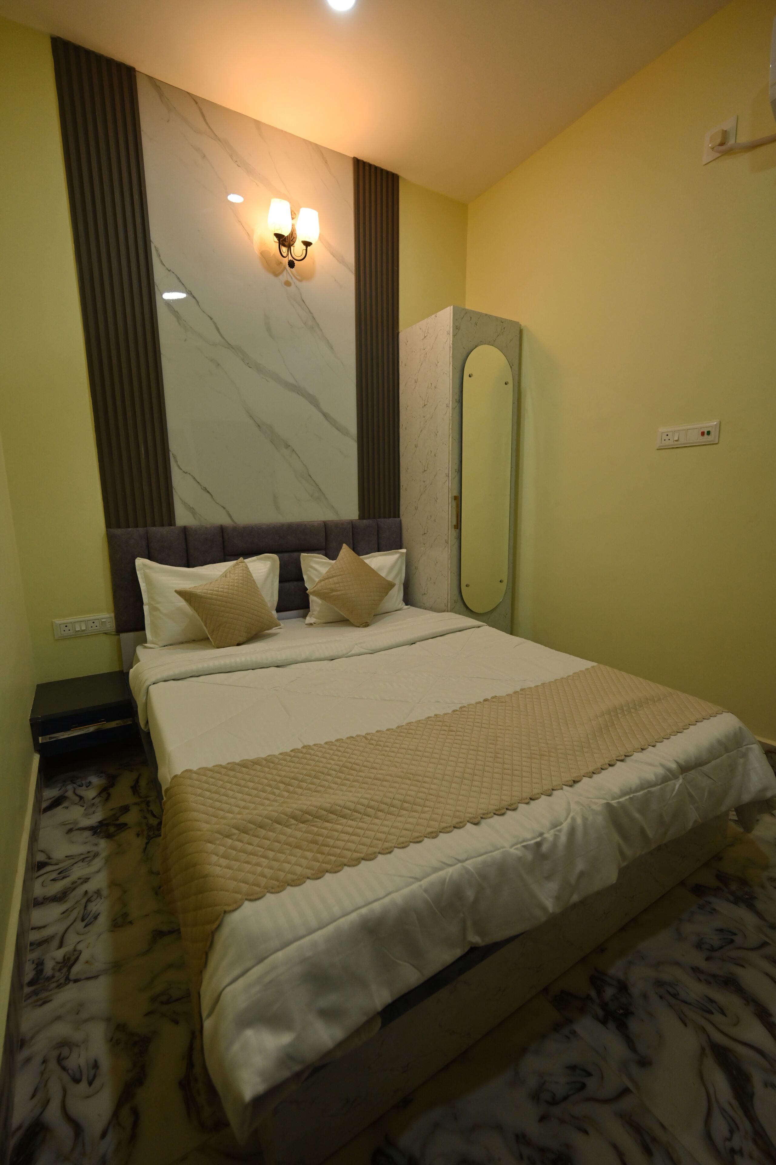 A hotel room with a bed, a headboard, a wall-mounted light fixture, a mirror, and a wall outlet.