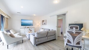 A modern living room with a grey sofa, white armchairs, a coffee table, and a TV. There is a desk with a computer and a chair. The room has white walls and a wooden floor. - Yawa at Yoolarai, 64 Yoolarai Cr - Ducted air, WiFi, Shared Pool (Nelson Bay)