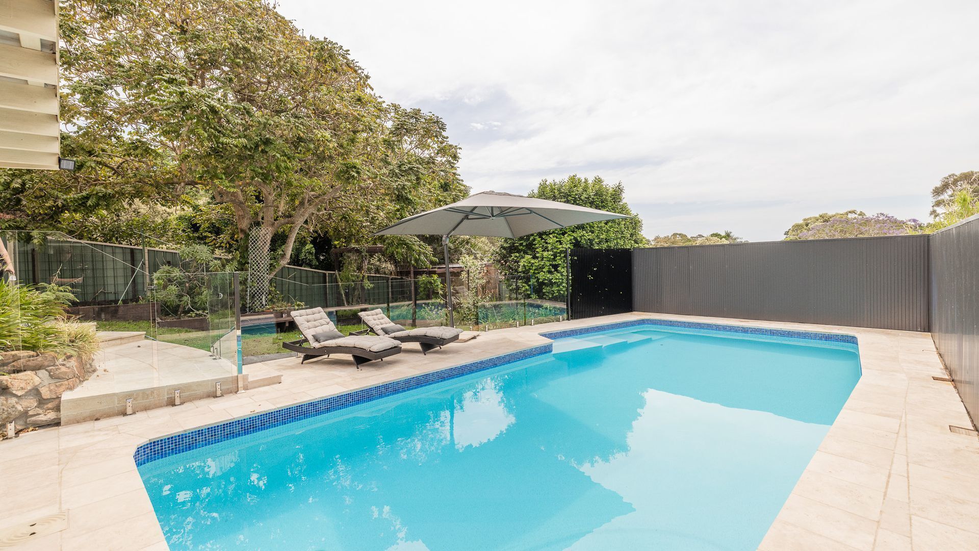 Ein rechteckiger Pool mit einer gefliesten Kante, umgeben von einer gepflasterten Liegefläche. Zwei Liegestühle mit Kissen und ein großer Sonnenschirm spenden Schatten. Das Pool-Areal ist durch einen Zaun begrenzt, im Hintergrund sind Bäume und Sträucher zu sehen.