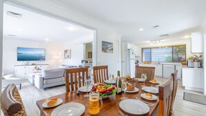 A modern dining area with a wooden table set for a meal, a kitchen in the background, and a living room with a sofa and TV. - Yawa at Yoolarai, 64 Yoolarai Cr - Ducted air, WiFi, Shared Pool (Nelson Bay)