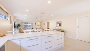 A modern kitchen with white cabinetry, a large island with drawers, and a dining area with white furniture. - Shelly's Bay Escape, 27c Dowling St - stylish with ducted air con and Wi-Fi (Nelson Bay)
