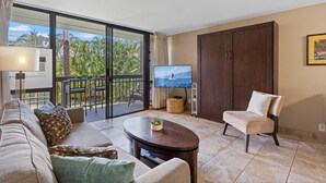 Sala de estar con sofá, sillón y una mesa de centro. Hay un televisor sobre un mueble bajo, una ventana con vista a palmeras y una puerta corrediza de vidrio que da a un balcón.
