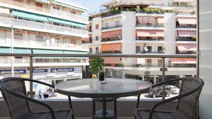 A balcony with wicker chairs and a glass table, overlooking a multi-story building with balconies and shops in the background. - Athenian Comfort Apartments (Zografos)