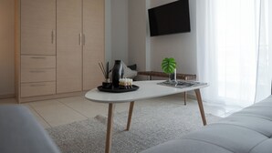 A modern living room with a white sofa, a coffee table with decorative items, a TV mounted on the wall, and a large window with sheer curtains. - Athenian Comfort Apartments (Zografos)