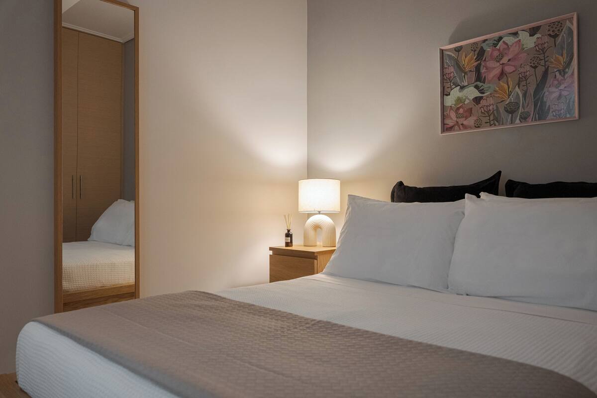 A neatly made bed with white linens, a bedside lamp, a wooden nightstand, and a mirror reflecting the room.
