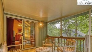 Una terraza de madera con mobiliario exterior, una puerta de cristal que da a una habitación interior y vistas a árboles.