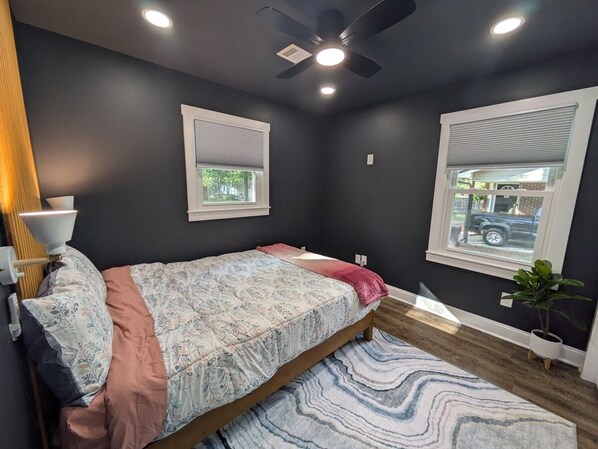 A bedroom with a bed, a ceiling fan, two windows with blinds, a lamp, and a potted plant.