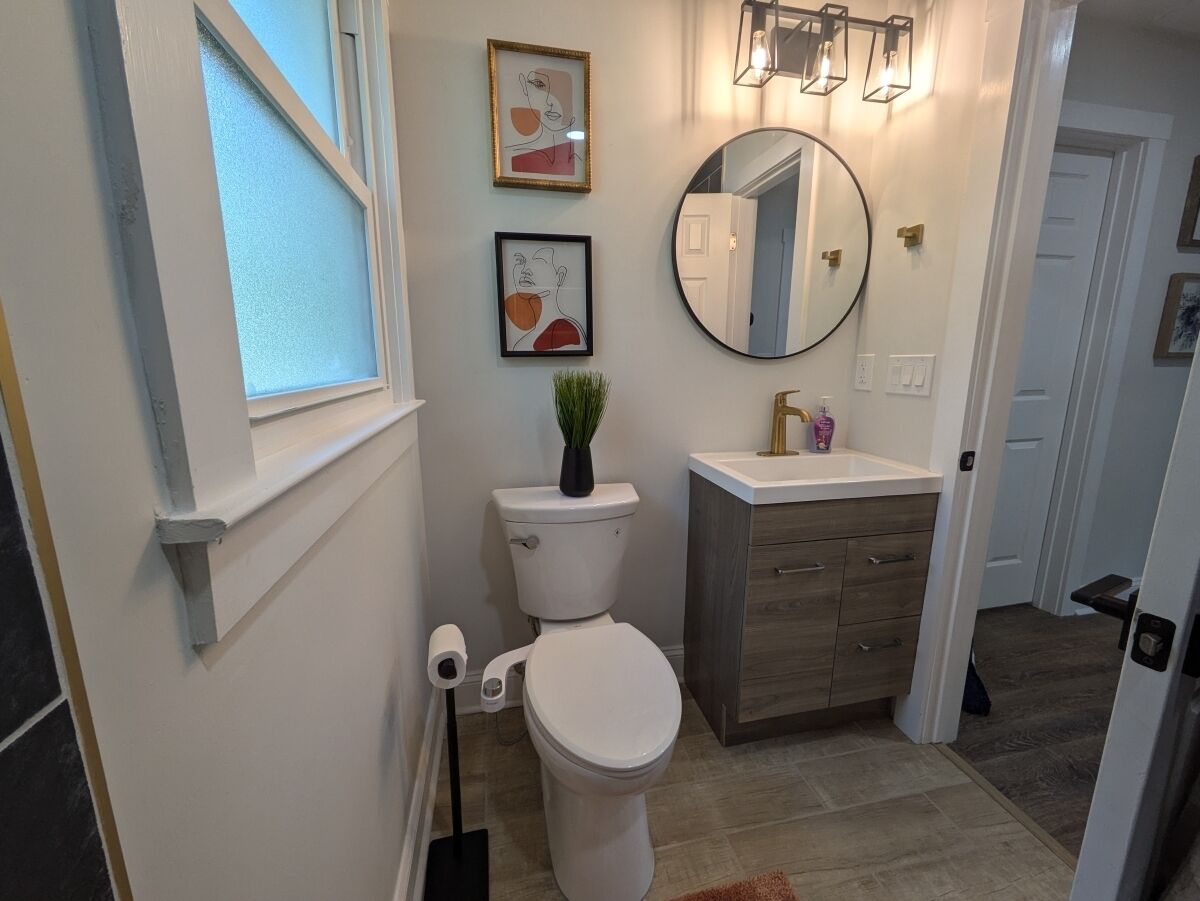 A modern bathroom with a toilet, sink, and mirror. There are framed artworks on the wall and a plant on the toilet.
