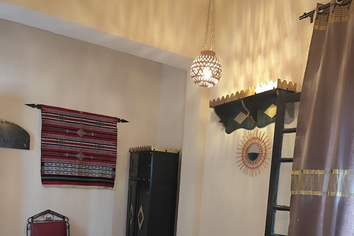 Quarto com duas camas, uma cadeira vermelha, um enfeite na parede, um candeeiro pendente e uma janela com cortina.
