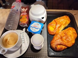 Tarjottimella on croissant, kuppi kahvia, pullo vettä, tetrapakki appelsiinimehua ja pussi sokeria.