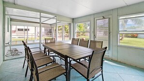 Une terrasse couverte avec une table et des chaises en bois, de grandes fenêtres et une vue sur l’extérieur.