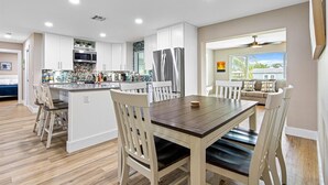 Une cuisine moderne avec des meubles blancs, un îlot central et un coin repas comprenant une table en bois et des chaises blanches.