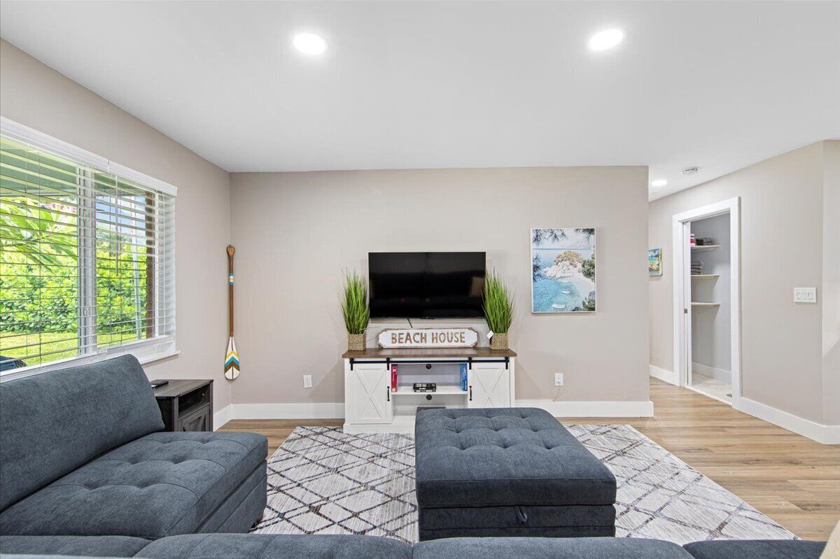 A modern living room with a grey sofa, ottoman, and a flat-screen TV on a wooden stand labeled "BEACH HOUSE".