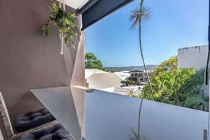 A balcony with a view of buildings and greenery, a hanging plant, and a cushioned seating area.